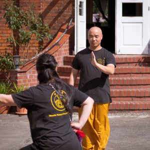 Meister Shi Yan Yao zeigt einer Kung-Fu-Schülerin eine Handtechnik im Innenhof der Zweigstelle Gollenberg. Im Hintergrund sind die Backsteinwand und der Treppenaufgang des historischen Gebäudes zu sehen.