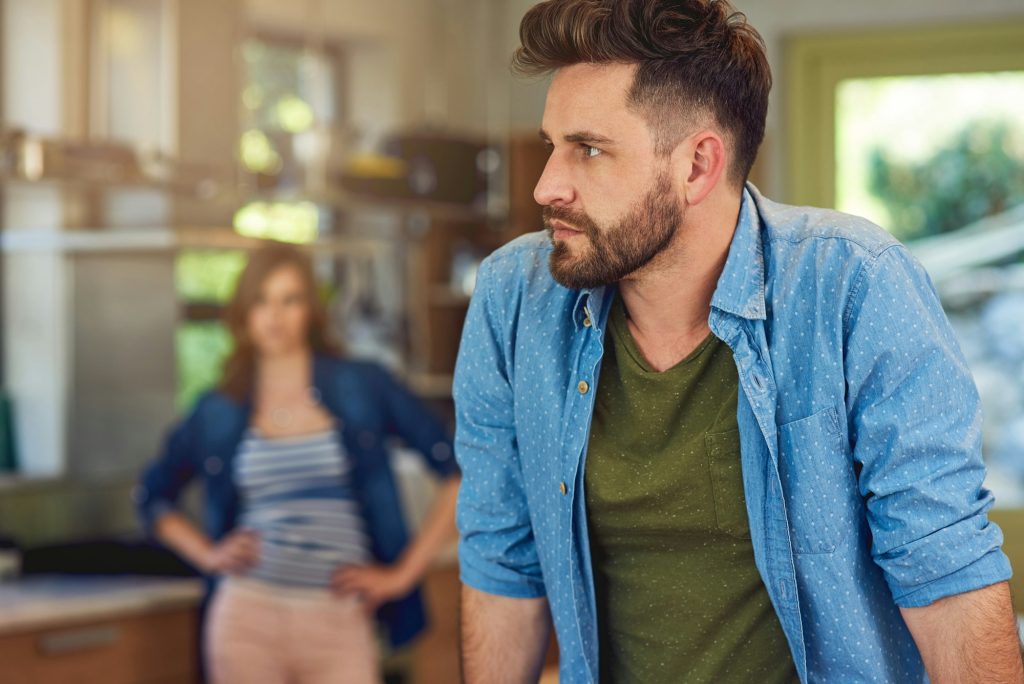 Shot of a young man looking upset after a fight with his wife who is standing in the background