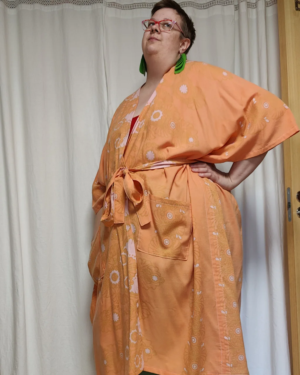 Medelålders tjock kvinna i en luftig kaftan Hemkram sydd av ett påslakan ljus orange bakgrund och ljusrosa blommor. Kaftanen är sydd av slow fashion-företaget Skamlös Design