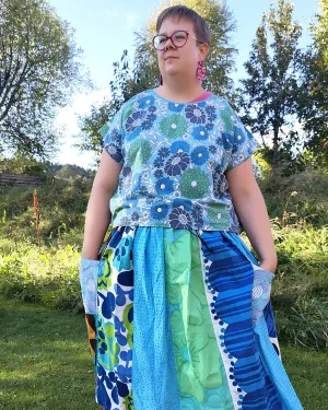 Medelålders korthårig tjock kvinna klädd i en kjol sydd i patchwork på längden i gröna, turkos och blå färger av återbrukad bomull. I bakgrunden en höstträdgård. Kjolen är sydd av slow fashion-företaget Skamlös Design
