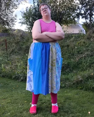 Medelålders korthårig tjock kvinna klädd i en kjol sydd i patchwork på längden i gröna, turkos och blå färger av återbrukad bomull. I bakgrunden en höstträdgård. Kjolen är sydd av slow fashion-företaget Skamlös Design