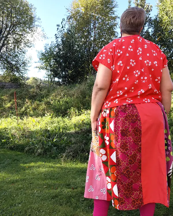 Medelålders korthårig tjock kvinna klädd i en kjol sydd i patchwork på längden i röda, rosa och vinröda nyanser av återbrukad bomull. I bakgrunden en höstträdgård. Kjolen är sydd av slow fashion-företaget Skamlös Design