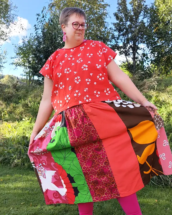 Medelålders korthårig tjock kvinna klädd i en kjol sydd i patchwork på längden i röda, rosa och vinröda nyanser av återbrukad bomull. I bakgrunden en höstträdgård. Kjolen är sydd av slow fashion-företaget Skamlös Design