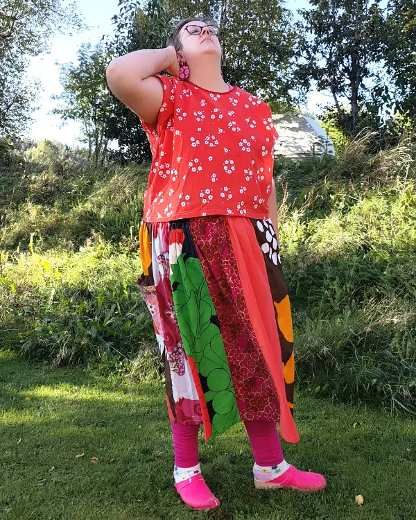 Medelålders korthårig tjock kvinna klädd i en kjol sydd i patchwork på längden i röda, rosa och vinröda nyanser av återbrukad bomull. I bakgrunden en höstträdgård. Kjolen är sydd av slow fashion-företaget Skamlös Design