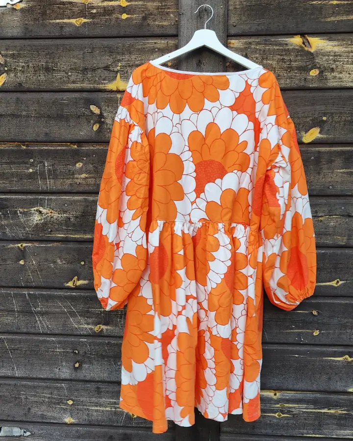 Klänning är sydd av ett påslakanstyg från IKEA med orange blommor och vita detaljer. Klänningen har långa ärmar, en avskärning vid midjan och en kjoldel med generös vidd och fickor. Sydd av Skamlös Design. Klänningen hänger mot en husvägg.