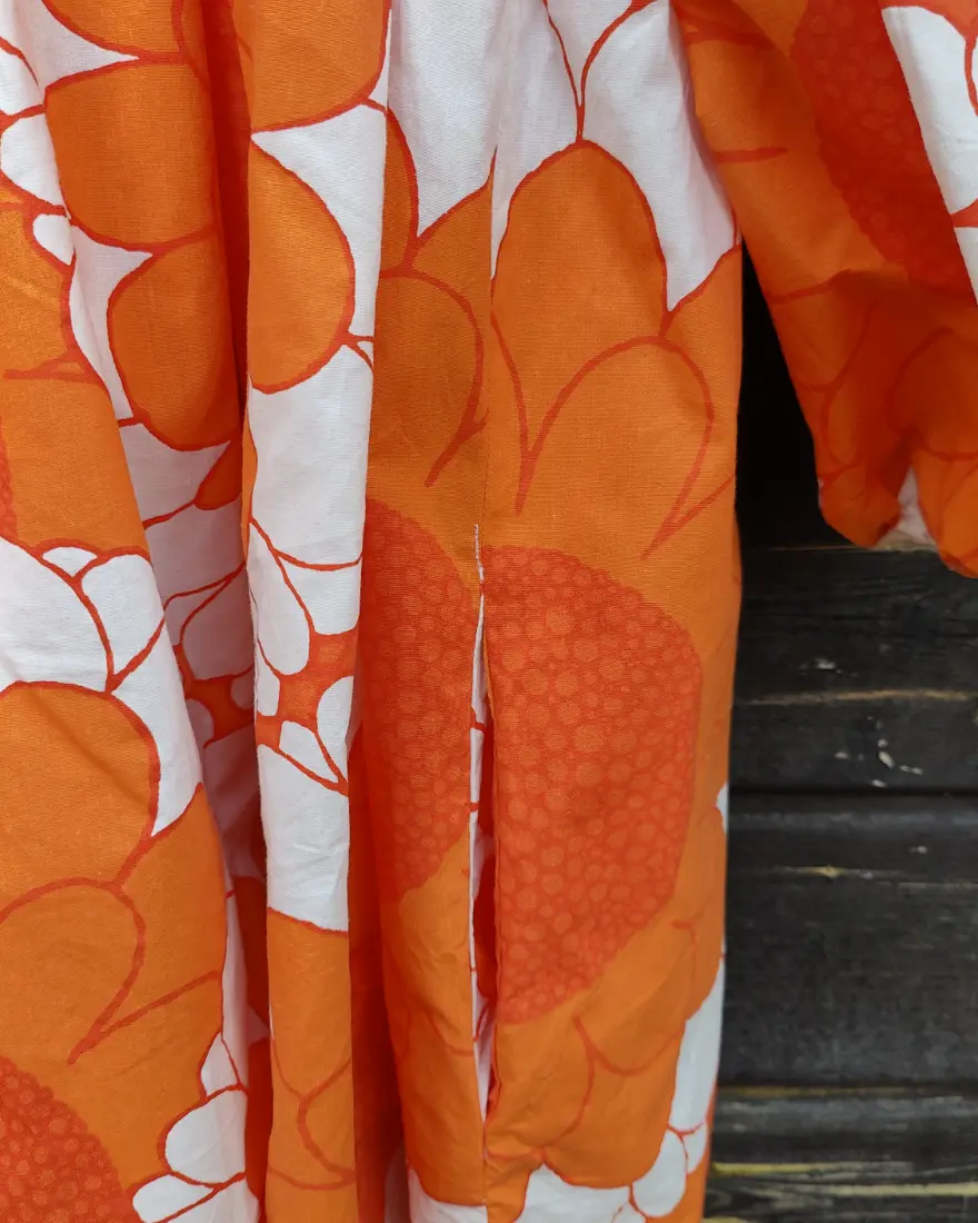 Klänning är sydd av ett påslakanstyg från IKEA med orange blommor och vita detaljer. Klänningen har långa ärmar, en avskärning vid midjan och en kjoldel med generös vidd och fickor. Sydd av Skamlös Design. Klänningen hänger mot en husvägg.