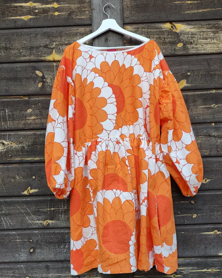 Klänning är sydd av ett påslakanstyg från IKEA med orange blommor och vita detaljer. Klänningen har långa ärmar, en avskärning vid midjan och en kjoldel med generös vidd och fickor. Sydd av Skamlös Design. Klänningen hänger mot en husvägg.