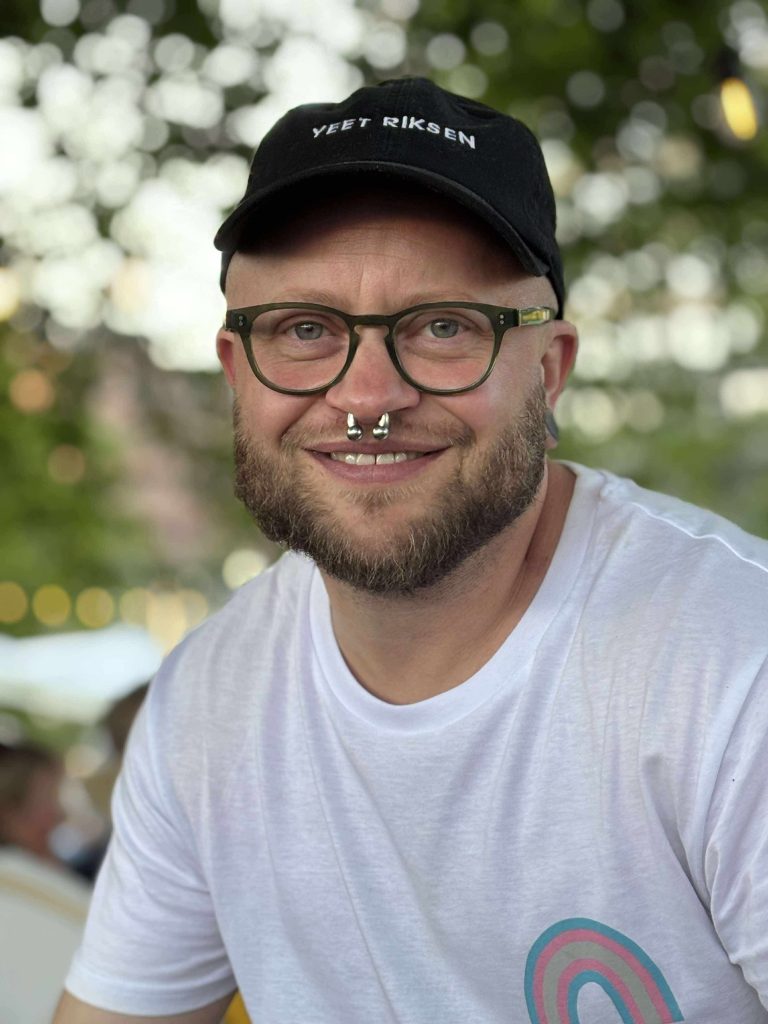 Portrettbilde av Luca, lys i huden, avrundede briller og kortklipt skjegg. Han har en sølvfarget septum-piercing i nesa, og har på seg en svart caps med teksten "yeet riksen" og en hvit t-skjorte med en trans regnbue på.