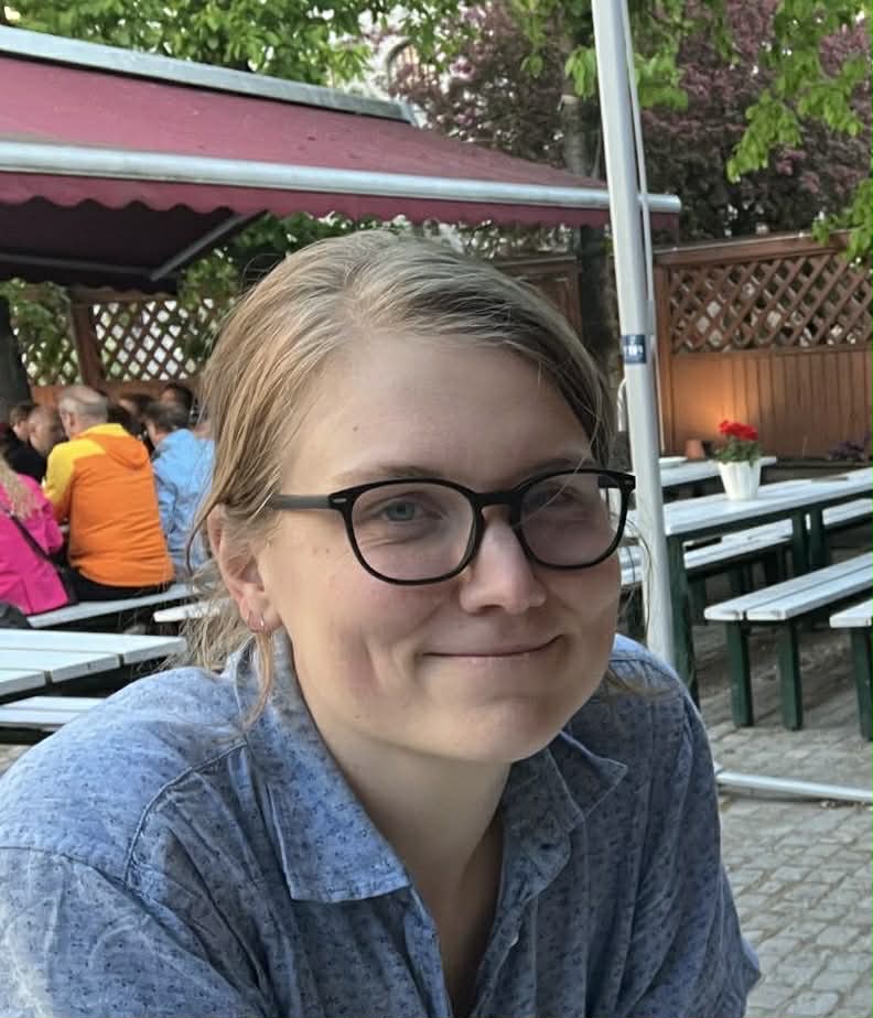 Bilde av Mar, hen er lys i huden med blondt hår trukket bak i en knute, svarte avrundede briller og en blå skjorte. De smiler med lukket munn til kamera. Bakgrunnen er en uteplass med flere bord med benker