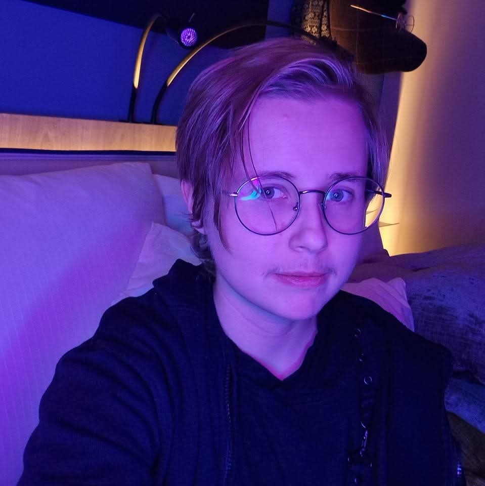 Portrettbilde av Lux, lys hud, kort blondt hår som er lengre foran, runde briller og svarte klær. Han sitter i et mørkt soverom med lilla lyssetting