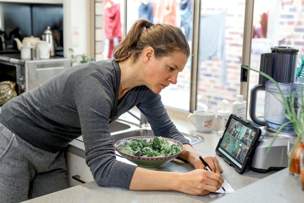 Woman in the kitchen watching zoom-meeting on tablet and writes notes