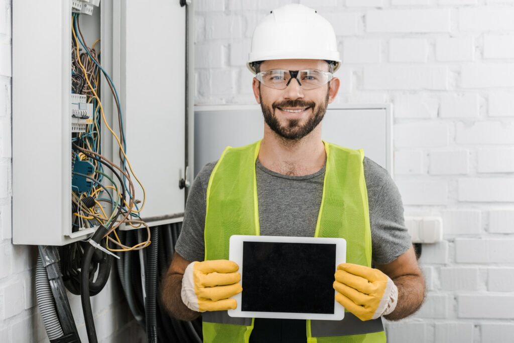 smiling handsome electrician showing tablet with blank screen near electrical box in corridor