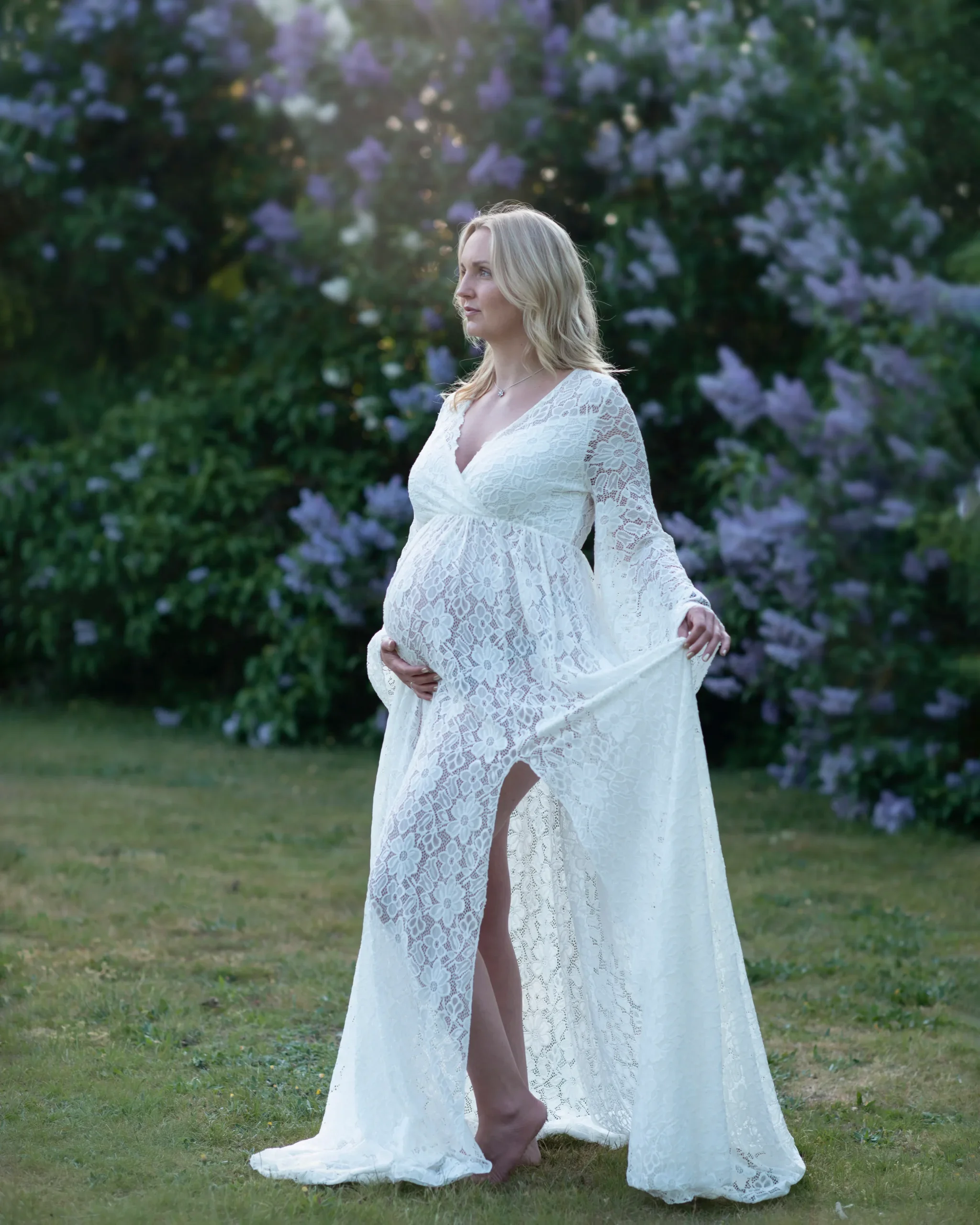 Gravidfotografering 5 Gravid kvinna med en vit klänning. I bakgrunden finns lila syrener. Gravidfotografering i Skåne