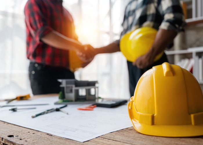 Yellow safety helmet on workplace desk with construction worker team hands shaking greeting start up plan new project contract in office at construction site, partnership and contractor concept.