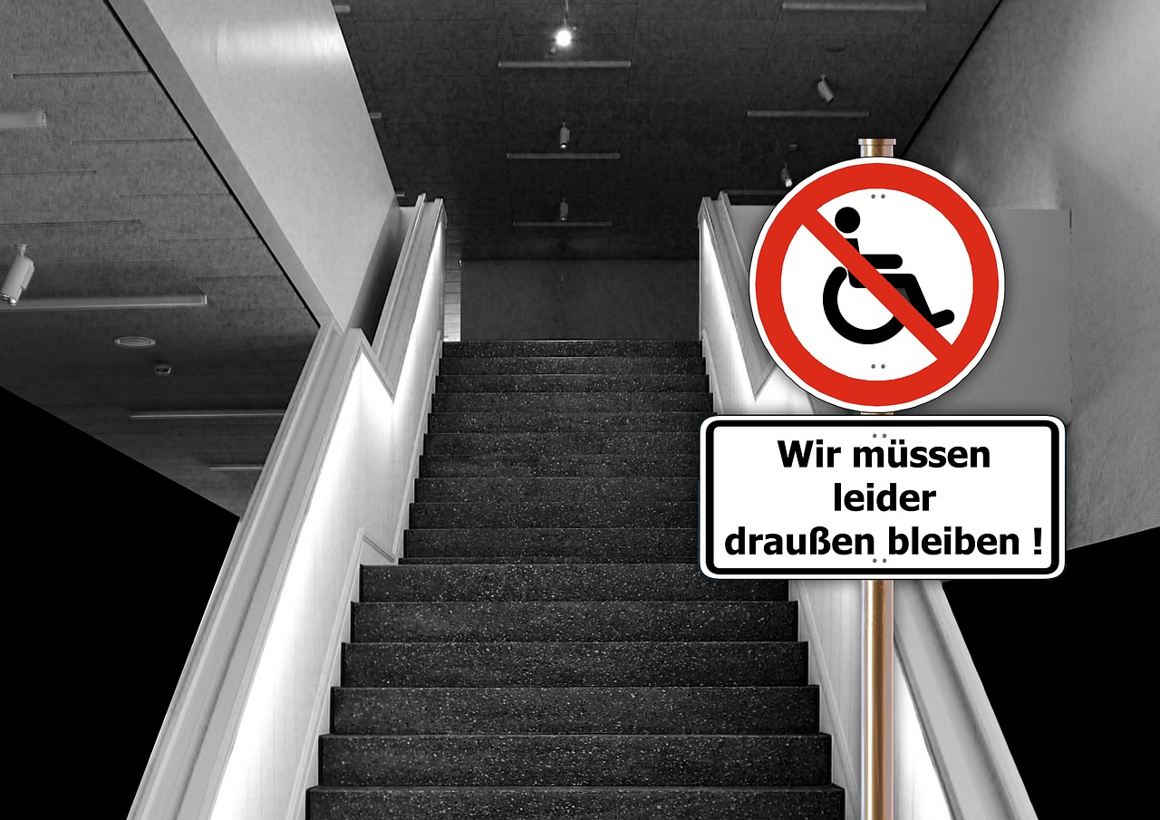 Eine Treppe. Davor steht ein Schild mit einem durchgestrichenen Rollstuhlsymbol. Darunter der Zusatztext: "Wir müssen leider draußen bleiben"