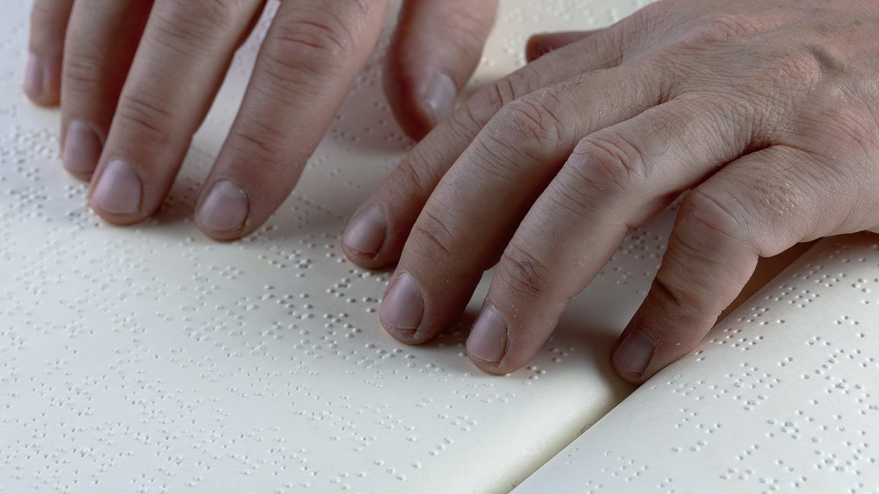 Zwei Hände, die in einem aufgeschlagenen Buch mit Braille Schrift lesen.