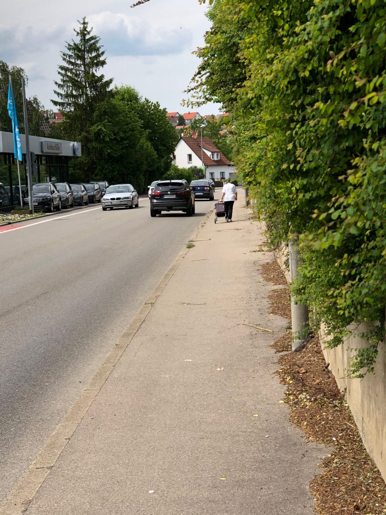 Eine Straße auf der in beiden Richtungen Fahrzeuge fahren. Rechts daneben ein Gehweg. Man sieht eine Frau mit Handwagen laufen. Rechts neben dem Gehweg eine Stützmauer und Sträucher, die leicht in den Gehweg hinein wachsen