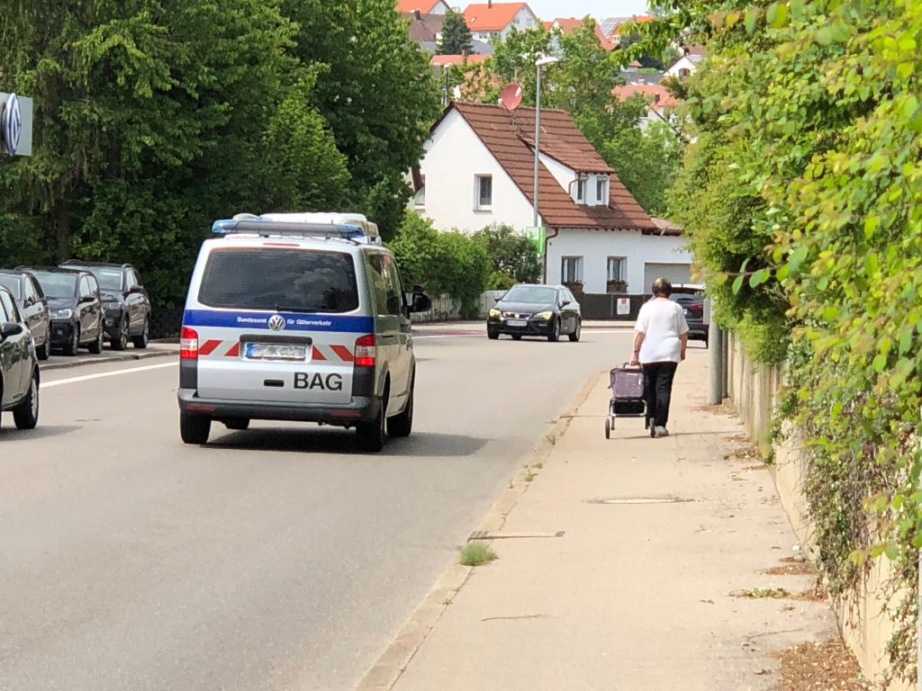 Eine Straße auf der in beiden Richtungen Fahrzeuge fahren. Rechts daneben ein Gehweg. Man sieht eine Frau mit Handwagen laufen. Rechts neben dem Gehweg eine Stützmauer und Sträucher, die leicht in den Gehweg hinein wachsen