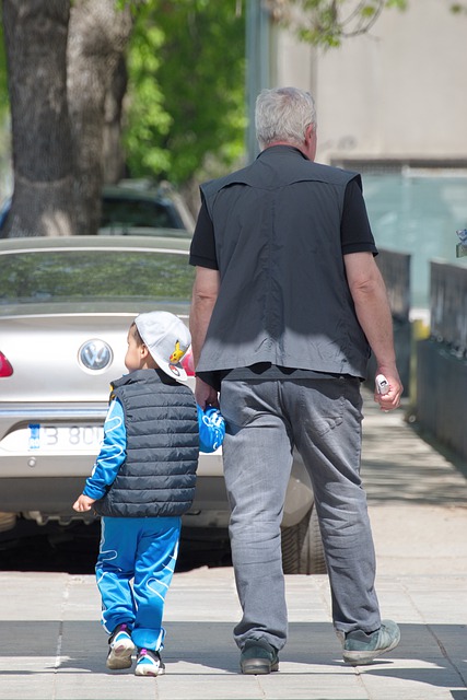 Ein Mann und ein Kind an seiner Hand von hinten. Beide laufen auf einem Gehweg. Auf dem Gehweg vor ihnen parkt ein Auto, so dass beide nur sehrt schlecht weiterlaufen können.