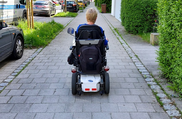 Ein breiter Gehweg mit gut berollbarem Pflasterbelag. Auf dem Gehweg fährt eine Frau in einem Elektrorollstuhl. Sie ist von hinten zu sehen.