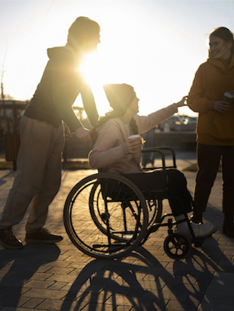 Drei Personen auf einem Platz, eine davon im Rollstuhl sitzend. Eine Person schiebt. Die drei Personen sehen vergnügt aus. Im Hintergrund ein Sonnenuntergang.