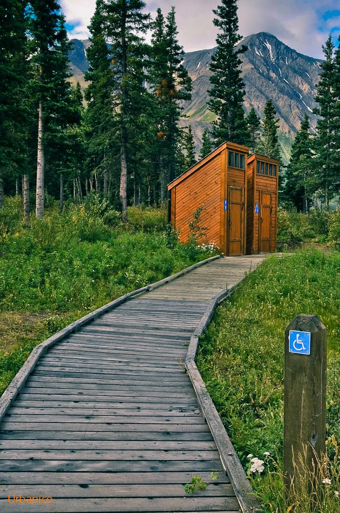 Zwei Klohäuschen aus Holz, eines davon als rollstuhlgerecht gekennzeichnet. Mitten in der Natur in Kanada am Kathleen Lake. Zu den Klohäuschen führt ein Holzplanken Weg mit seitlicher Aufkantung durch mit niederen Büschen und Wiese bewachsene Natur. Im Hintergrund Nadelbäume und ein Hoher Berg mit Resten von Schnee.