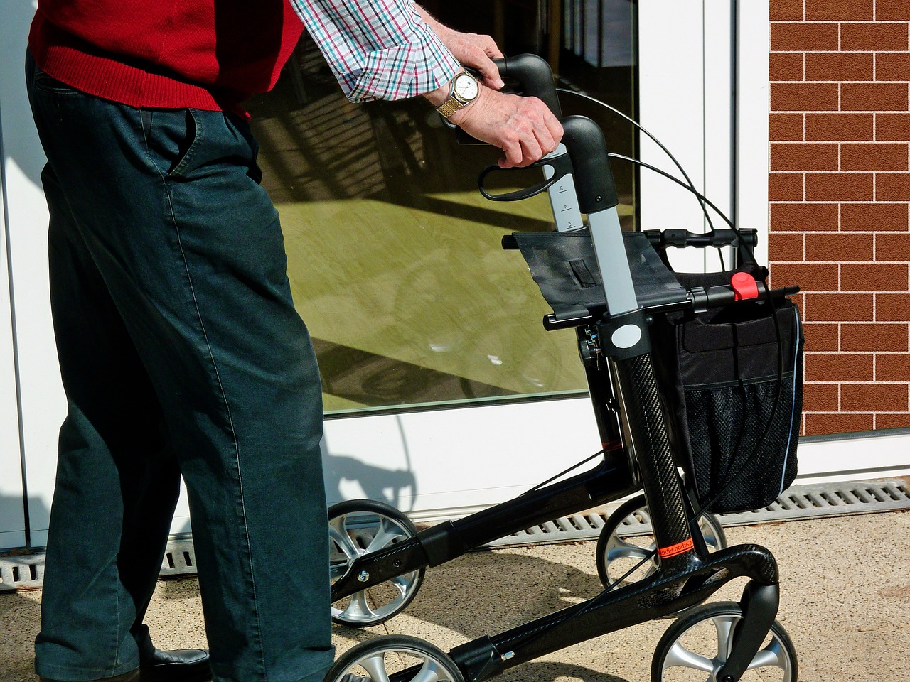 Ein Mann mit Rollator. Von dem Mann sieht man nur den Ausschnitt von den Händen, die Griffe des Rollators fassen, bis oberhalb der Füße. Er läuft auf einer gut berollbaren Fläche an einer Hauswand mit Terrassentür entlang.