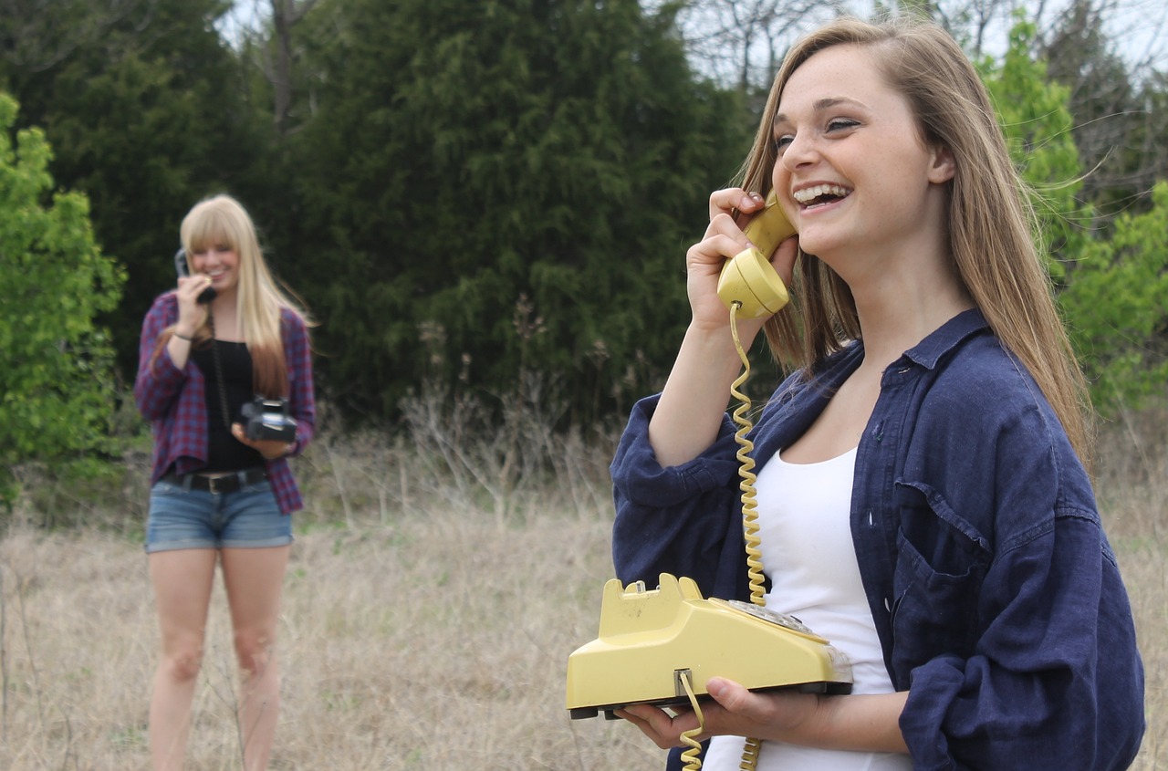 Zwei junge Frauen stehen auf einer Wiese und haben jeweils ein altes Wählscheibentelefon in der einen Hand. In der anderen Hand den Telefonhörer ans Ohr gehalten. Sie stehen auf seiner Wiese, die eine weiter vorne, die andere weiter hinten.