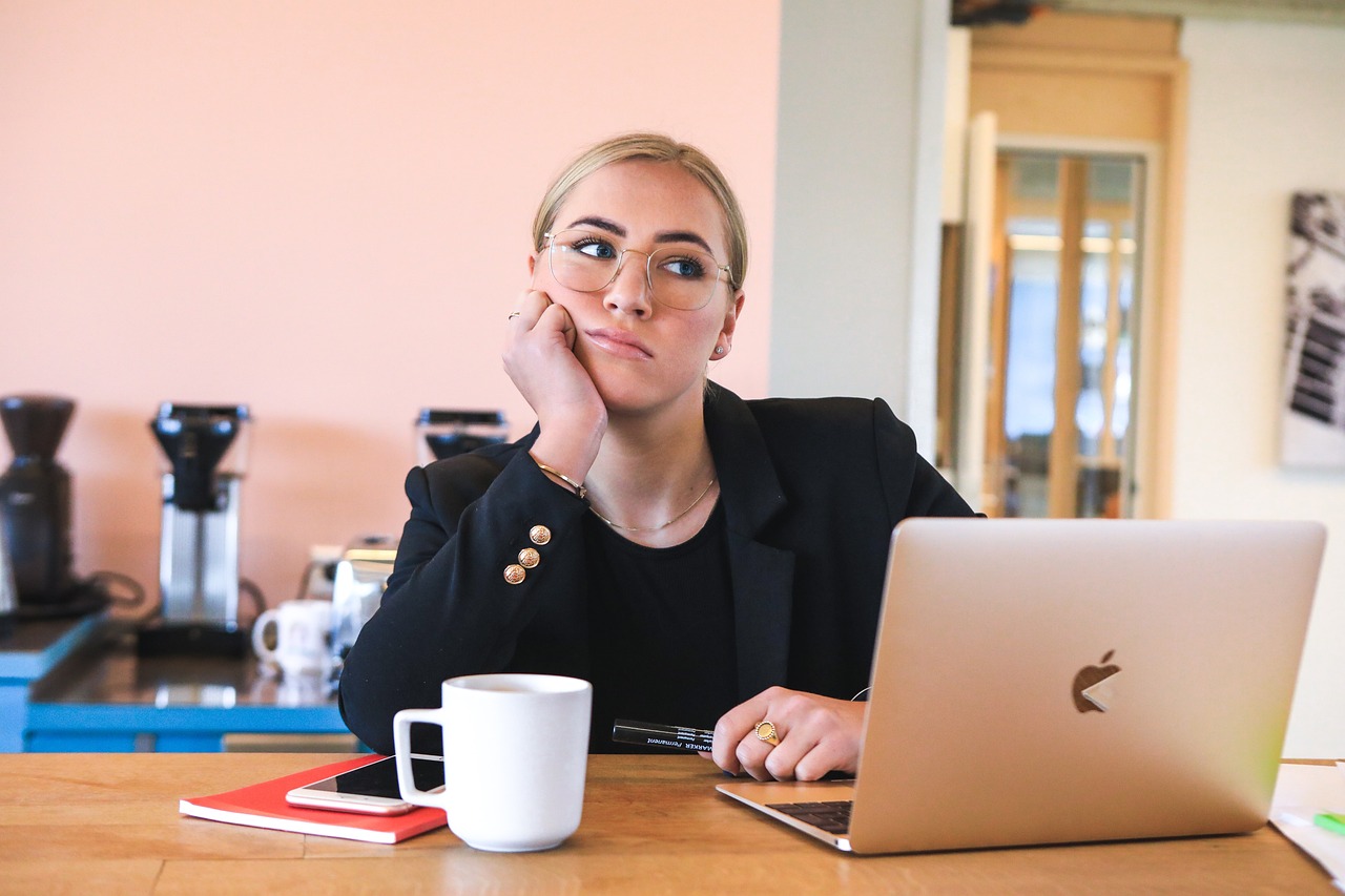 Frau, die am Tisch vor aufgeklapptem Laptop sitz, den Kopf auf die Hand gestützt. Sie schaut müde in die Weite. Neben dem Laptop steht eine weiße Kaffeetasse, daneben liegt ein Notizbuch und darauf ein Handy. Im Hintergrund unscharf, Kaffeemaschinen.