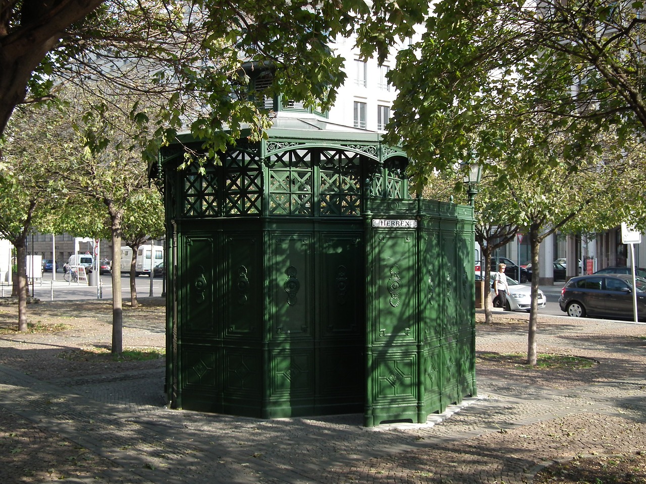 Alte öffentliche Toilette in Berlin am Gendarmenmarkt aus grün lackiertem Gusseisen unter Bäumen.