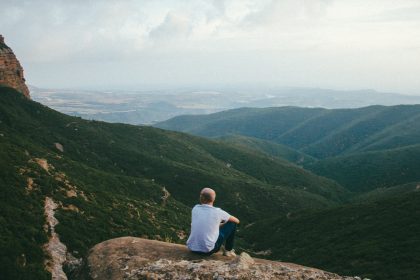 person sitting on the edge of a cliff over looking mountains during daytime