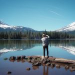 woman in white long sleeve shirt and blue denim jeans standing on brown log near lake