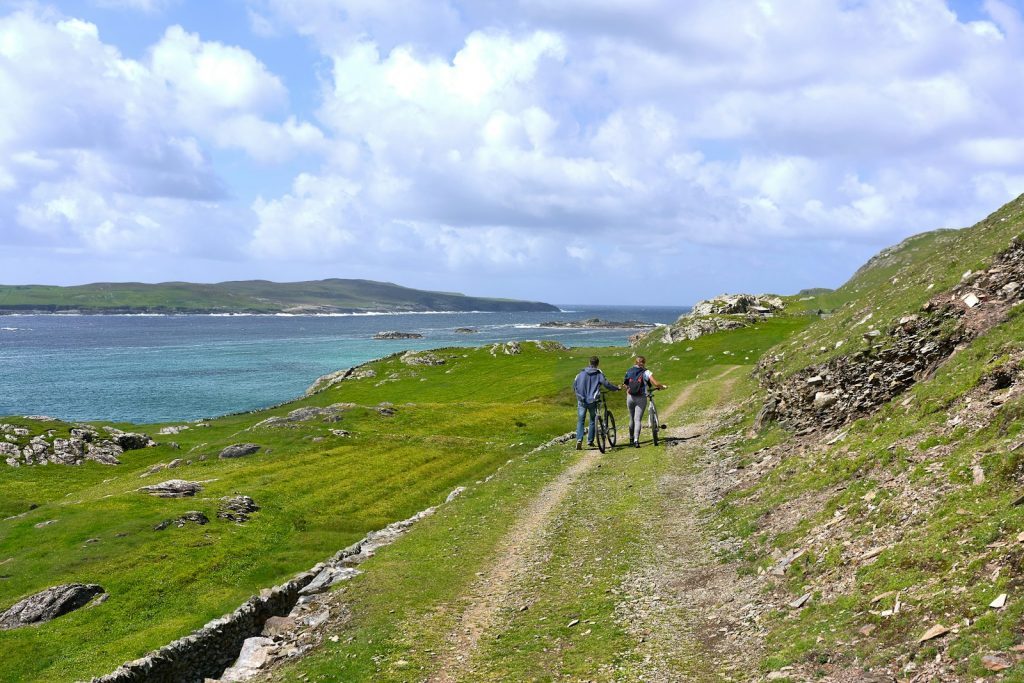 two people walking together with bikes in green field viewing mountain and blue sea under white and blue sky during daytime The Wild Atlantic Way Exploring the Open Road