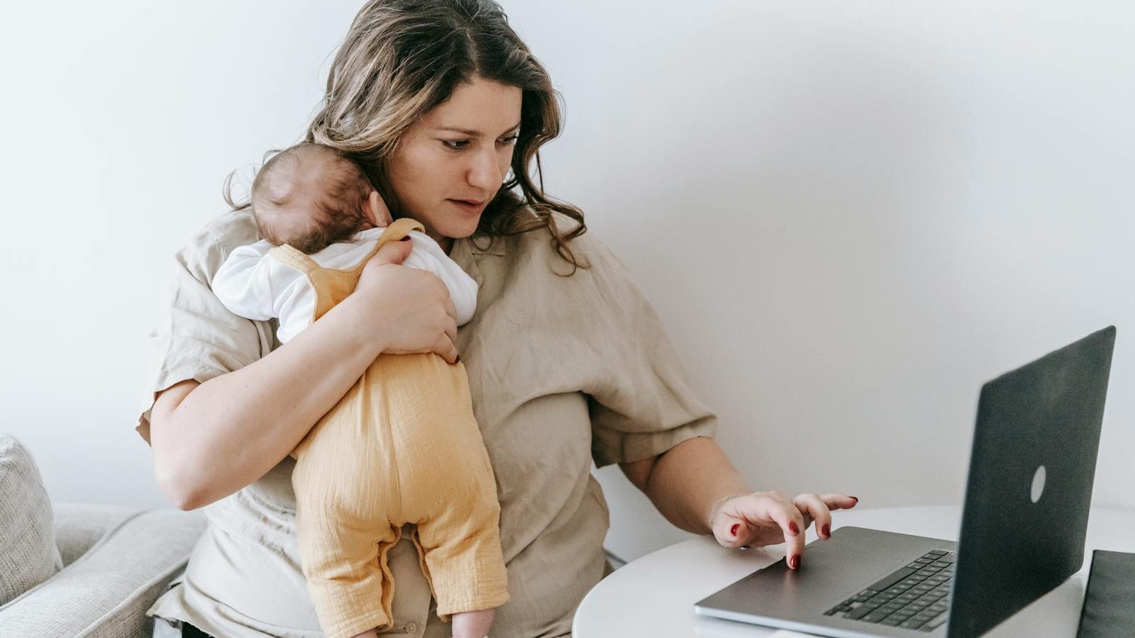 Concentrated young female freelancer embracing newborn while sitting at table and working remotely on laptop at home. 10 Life-Saving Parenting Hacks for Busy Moms