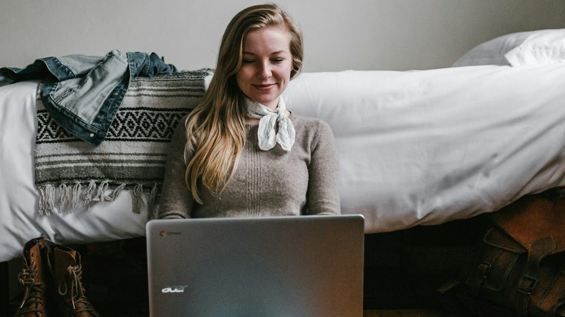 woman sitting beside a bed while using a laptop