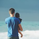 man wears blue crew-neck t-shirt holding toddler wears black hooded jacket near ocean under blue sky at daytime