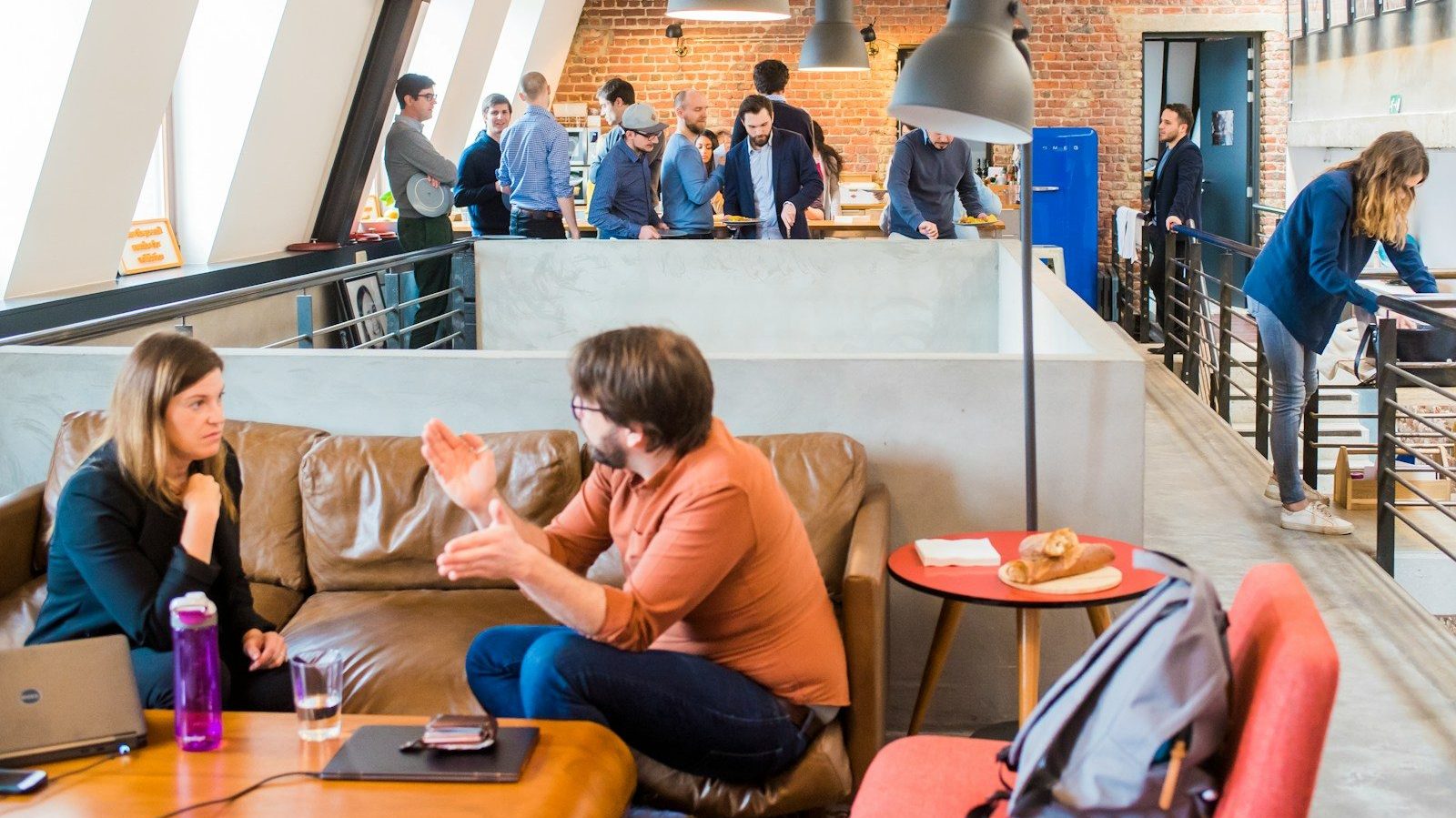 man in orange dress shirt talking and sitting in front of woman on brown sofa Workplace Openness Talking Money With Your Team