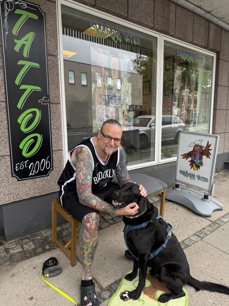 Tattooed man in a Brooklyn jersey sits on a bench outside a tattoo parlor, petting a large black dog; storefront window displays tattoo art panels. steeleye tattoo danmerks bedste tatovør odense svendborg dalum bolbro kerteminde Brian tattoo steeleye tattoo