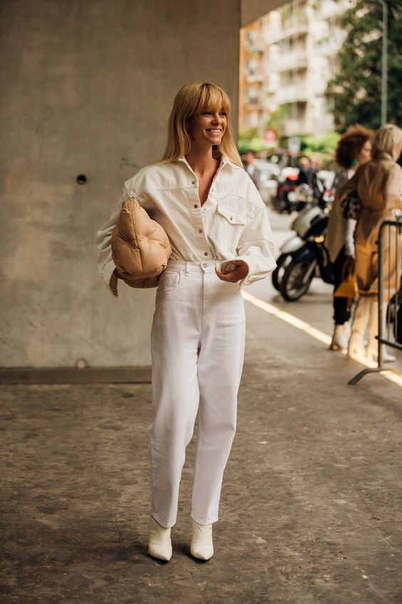 En kvinna i hel vit outfit. Vita jeans och vit jeansskjorta instoppat med en vääska i sidan som hon håller i. 