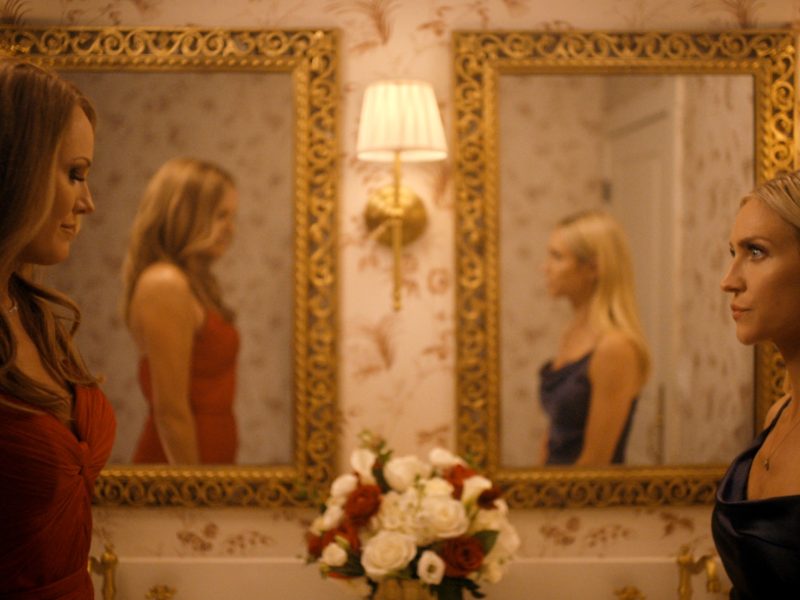 "Two women in elegant dresses stand facing each other in a warmly lit, ornate bathroom with floral wallpaper, gold-framed mirrors, wall lamps, and a vase of flowers on the counter, their reflections visible in the mirrors."