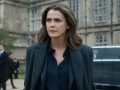 "A woman with wavy brown hair in a dark coat stands outside a grand historic building, looking serious. There is a black car and security personnel in the background, suggesting a formal or official environment."