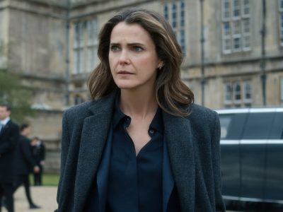 "A woman with wavy brown hair in a dark coat stands outside a grand historic building, looking serious. There is a black car and security personnel in the background, suggesting a formal or official environment."