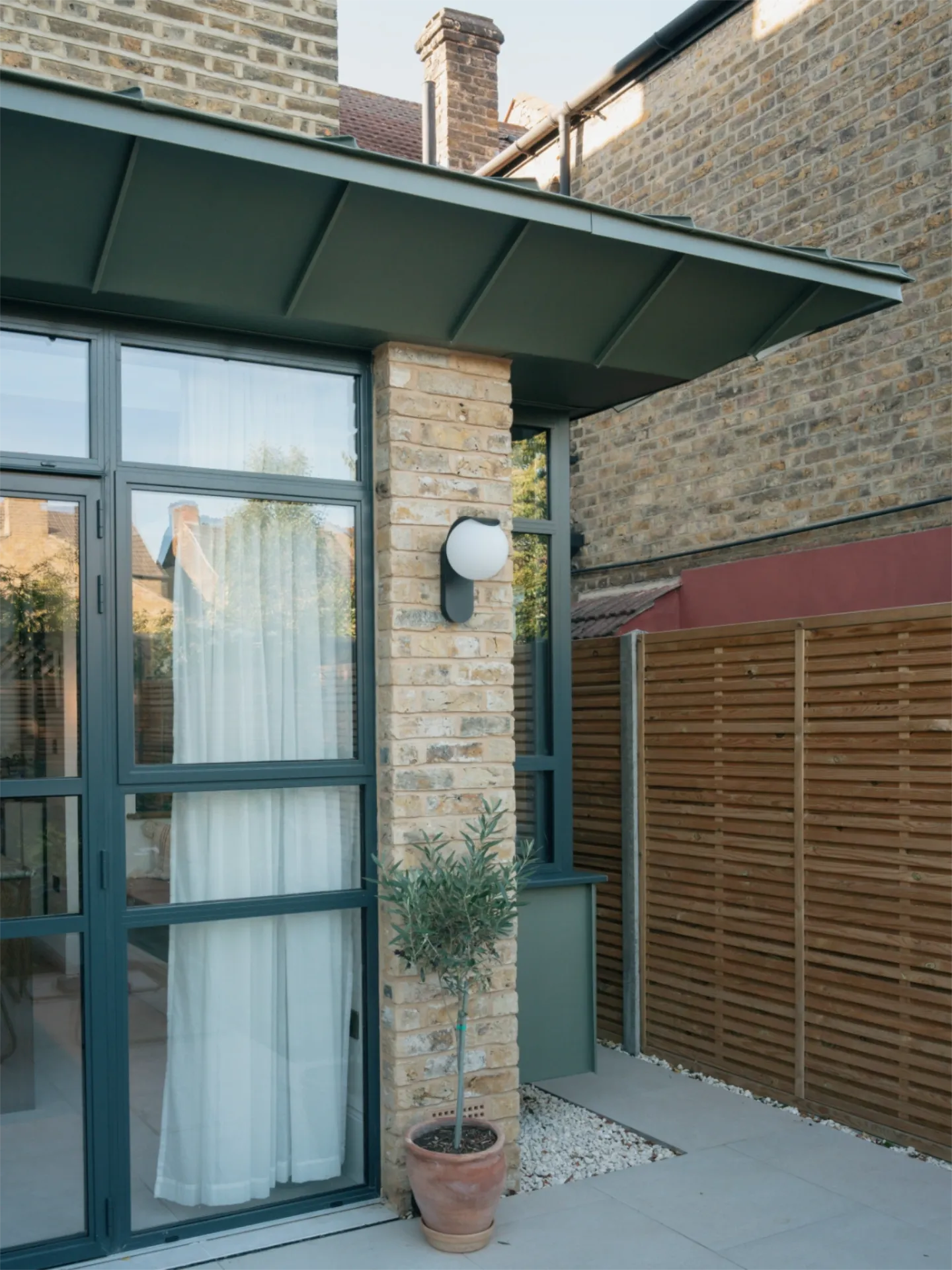 Close-up of a modern rear extension at Tooting House, showing a green standing-seam zinc canopy, reclaimed yellow London stock brickwork, a globe wall light, and slim-profile aluminium glazing.