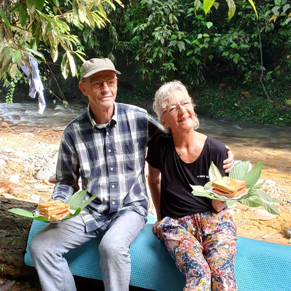 Elderly couple sitting on a blue mat by a stream in the jungle, smiling and holding sandwiches wrapped in green leaves during a break on their trek