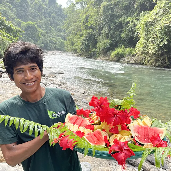 guide smiling and holding a tray of vibrant watermelon slices and tropical flowers, with a serene river and lush jungle in the background