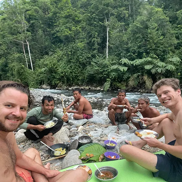 happy group sitting on rocks by a river in the jungle, enjoying a shared meal with a backdrop of lush tropical forest