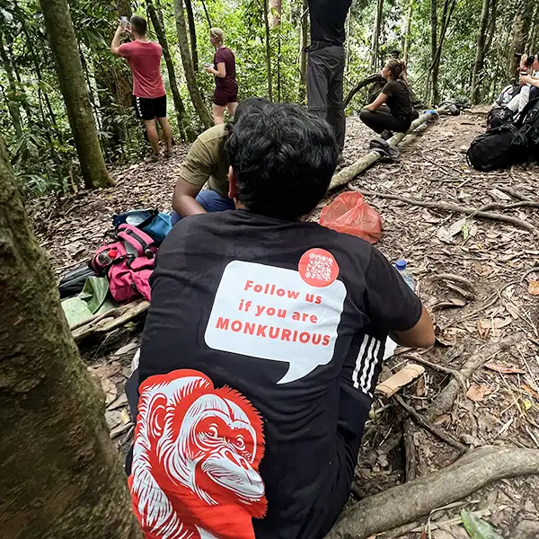 Resting hikers in the jungle with a guide's shirt humorously inviting onlookers to follow if they are "MONKURIOUS", featuring a playful orangutan face.