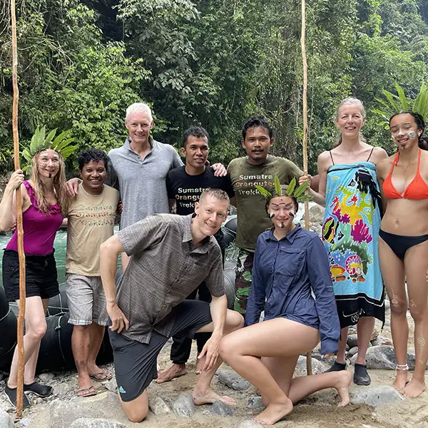 A diverse group of hikers smiling at the camera in a lush forest setting, some holding walking sticks and backpacks, after a trekking adventure.