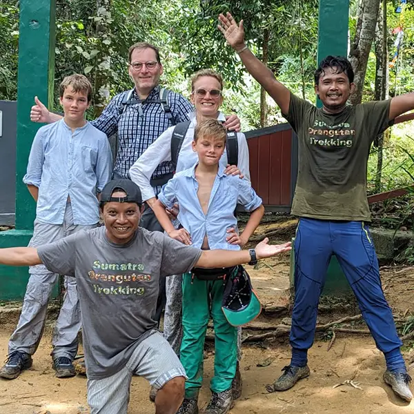 A family and trekking guides pose with thumbs up and cheerful expressions at a trailhead, ready for an adventure in the jungle.