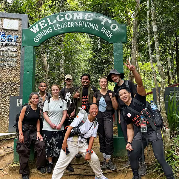 A group of trekkers with beaming smiles under the 'Welcome to Gunung Leuser National Park' sign, ready to explore the wilderness.